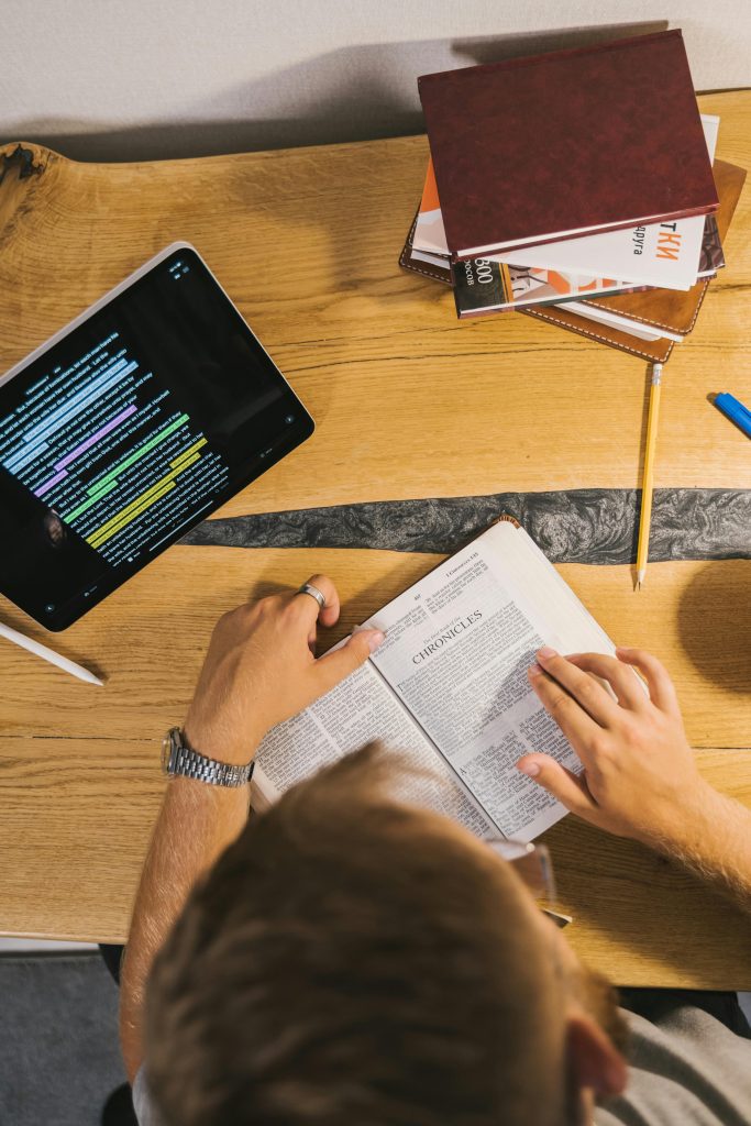 Person reading a Bible at a desk with a tablet and notebooks, emphasizing study and contemplation.