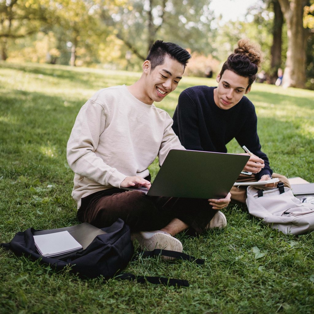 Full length content ethnic male students in casual outfits browsing laptop while sitting on meadow with copybooks and preparing for exam in green park