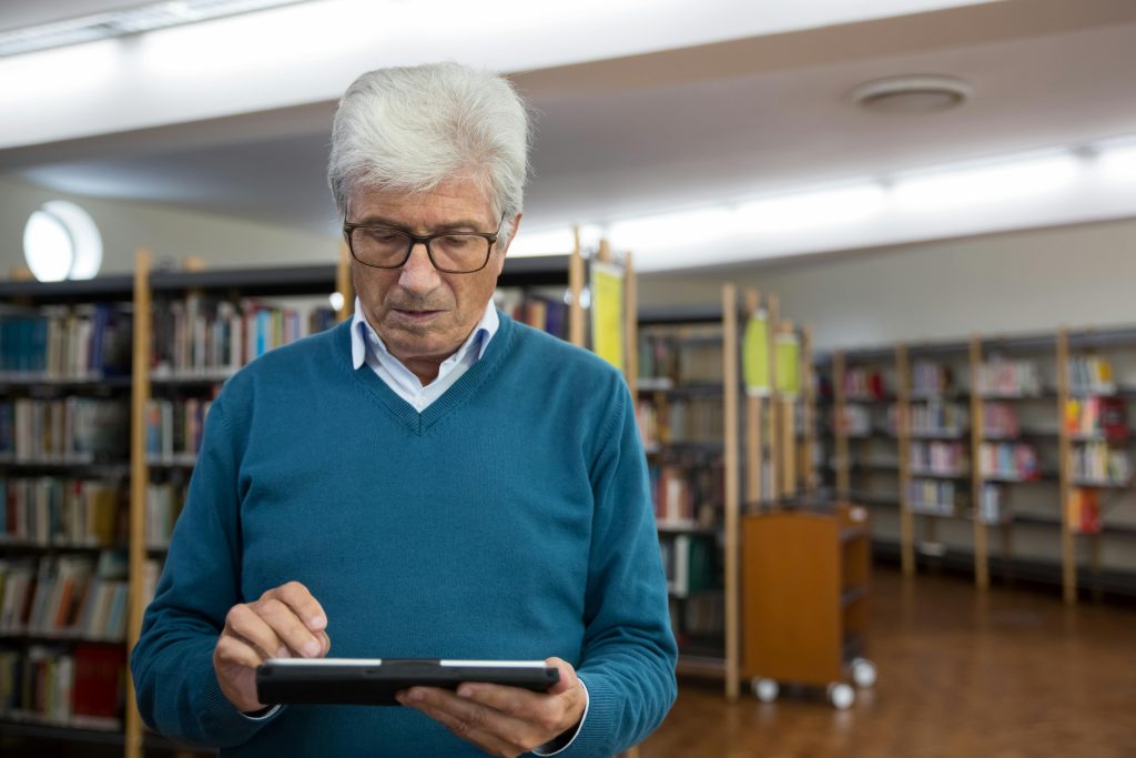 Elderly man engaging with technology on a tablet in a library setting.